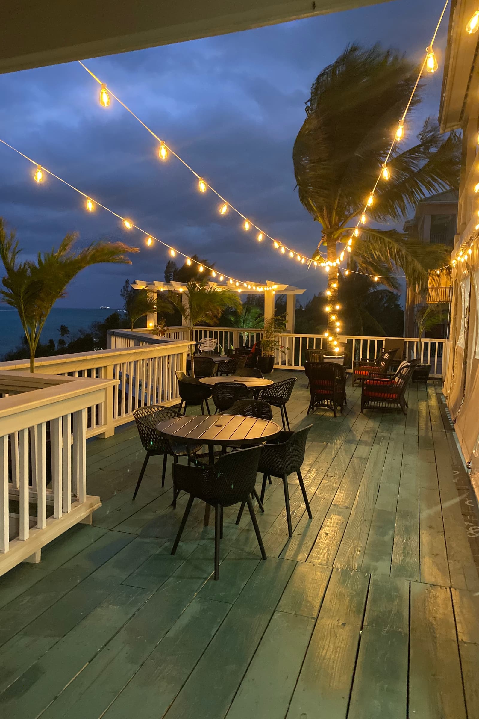 The deck at blue hour, filament lights strung overhead above bistro tables and the darkening sea